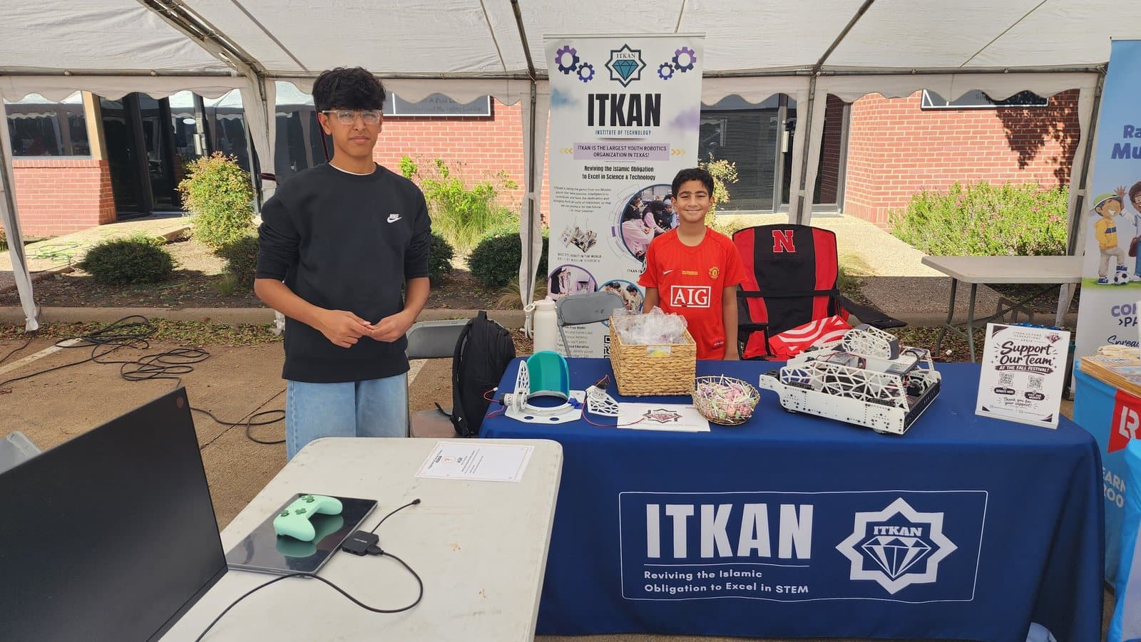 Two team members at the ITKAN table under a white tent at LCIC Fall Fest, their robot on display