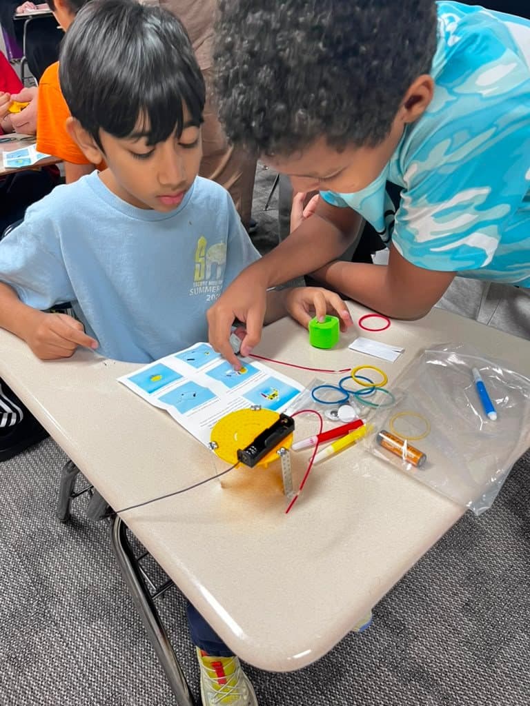 Two children assemble a scribble bot with a battery pack and markers