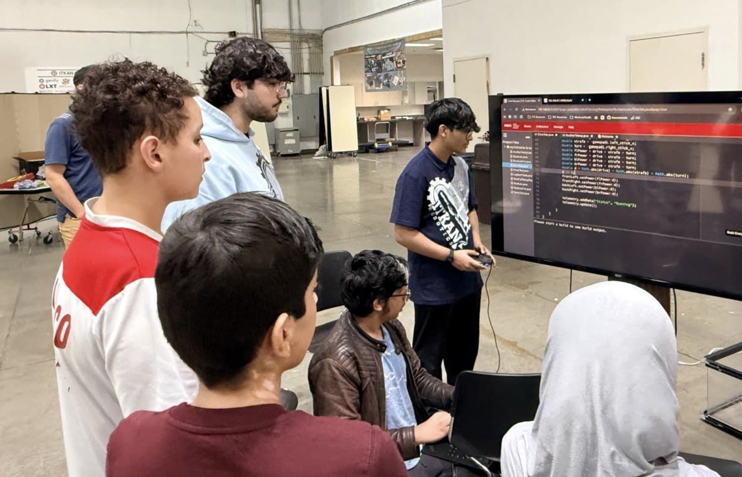 ITKAN students watch live teleop robot code on a large screen at the Innovation Hub while a student tests it with a controller