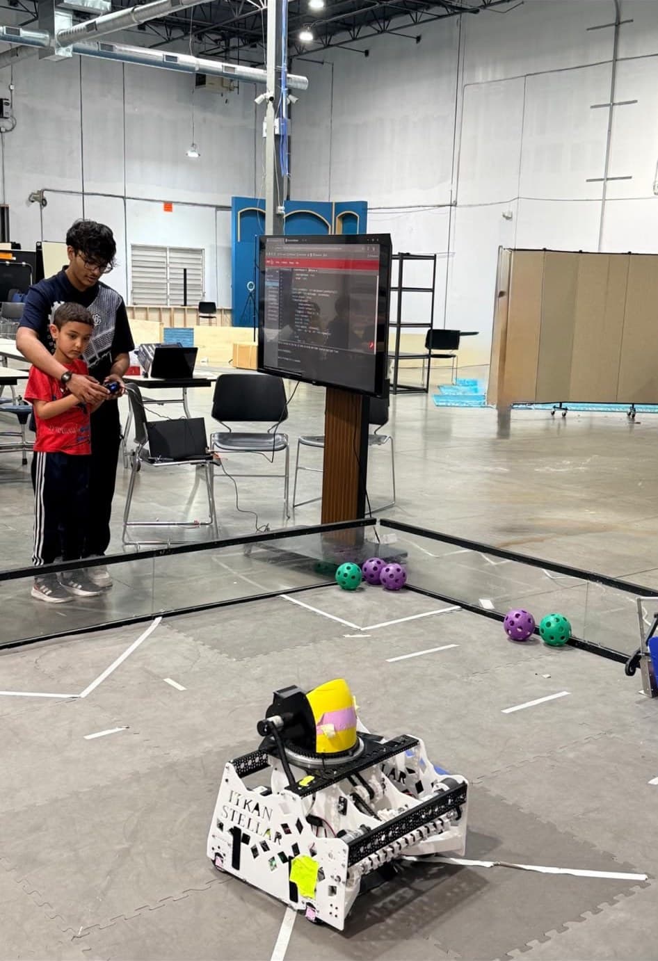 An ITKAN student mentor helps a young student drive the ITKAN Stellar robot on the practice field at the Innovation Hub