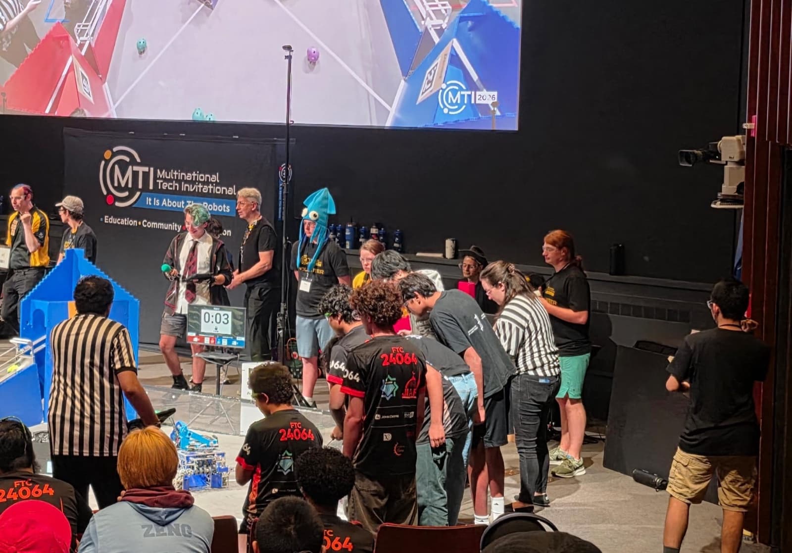 Teams crowd the MTI competition floor beneath a large Multinational Tech Invitational banner as a match clock counts down