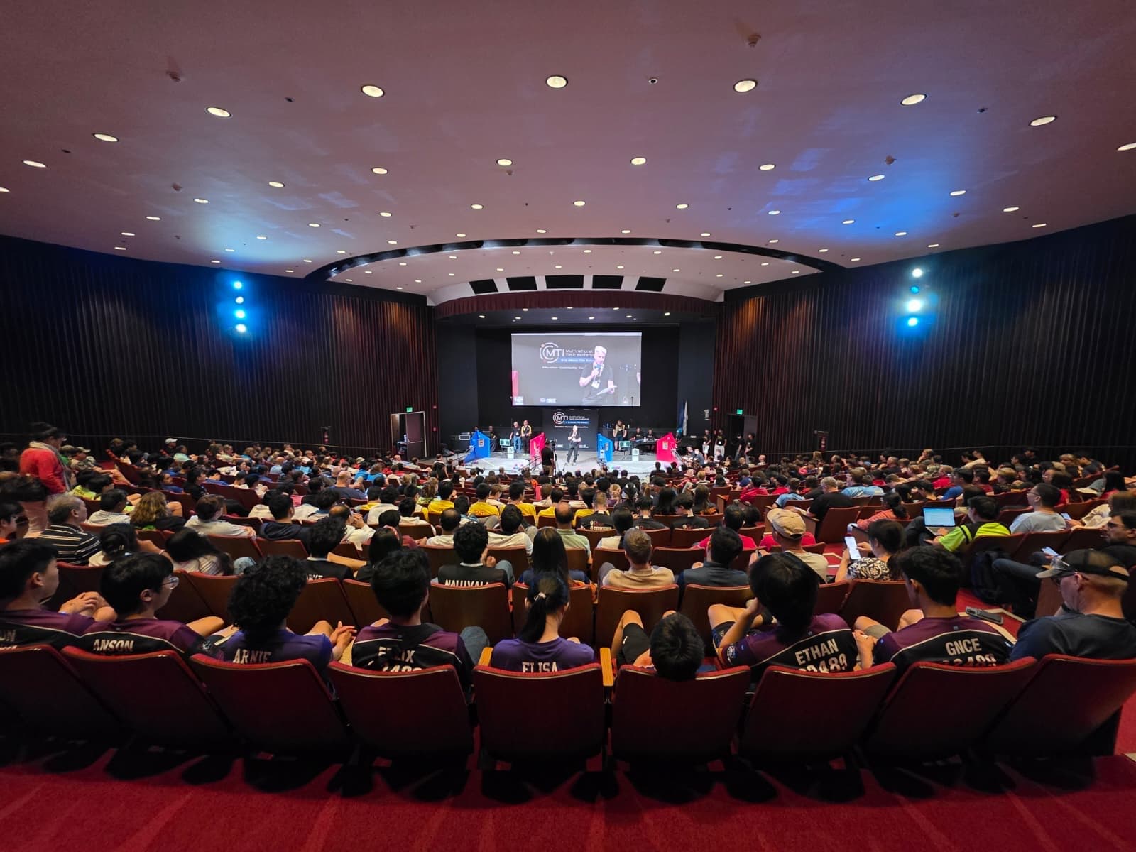A packed auditorium of teams and spectators faces the stage during a Multinational Tech Invitational ceremony