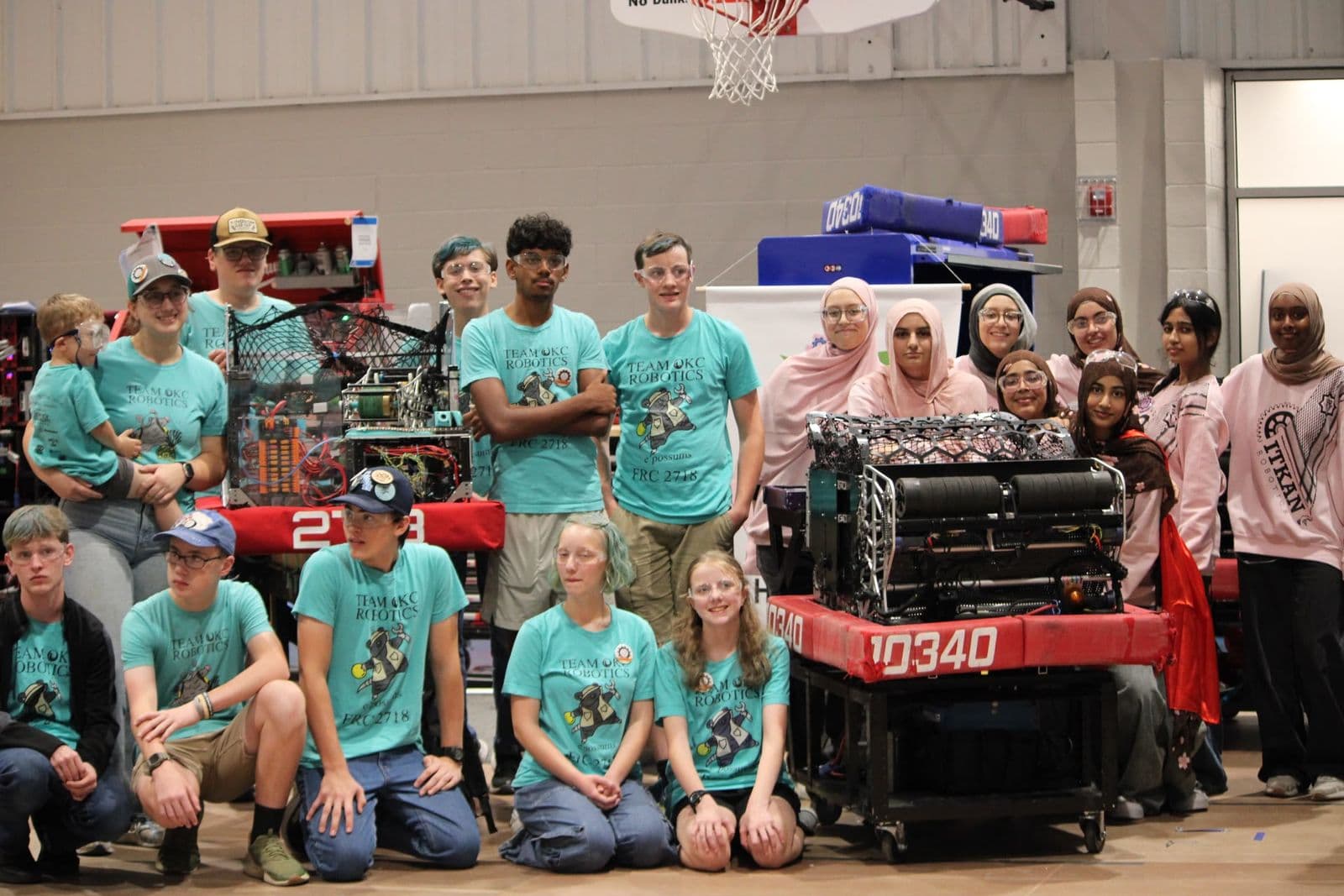 ITKAN Girls FRC Team 10340 in pink ITKAN hoodies and Team OKC Robotics members in teal shirts, posed with both teams' robots