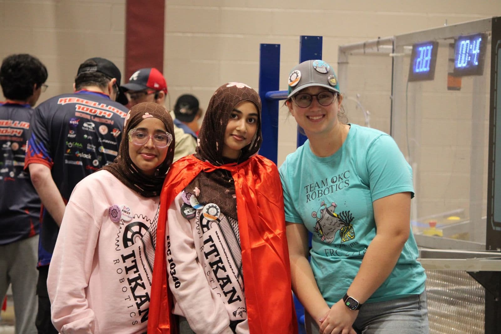 Two ITKAN Girls members in pink hoodies, one wearing a red cape, smile beside a Team OKC Robotics member in a teal shirt