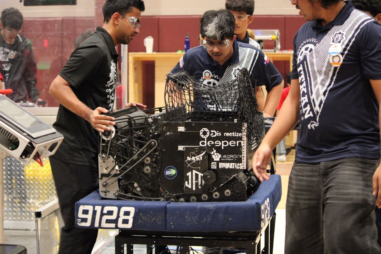 Team 9128 members in safety glasses gather around their robot on its cart