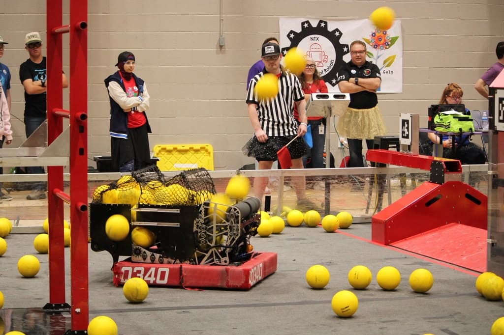 The 10340 robot on the field among yellow game pieces, with a referee and volunteers watching
