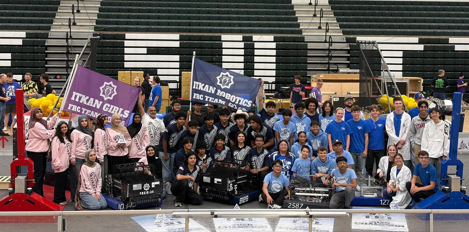 ITKAN Robotics 9128 and ITKAN Girls 10340 stand together on the TRI competition floor, holding their team banners and robots