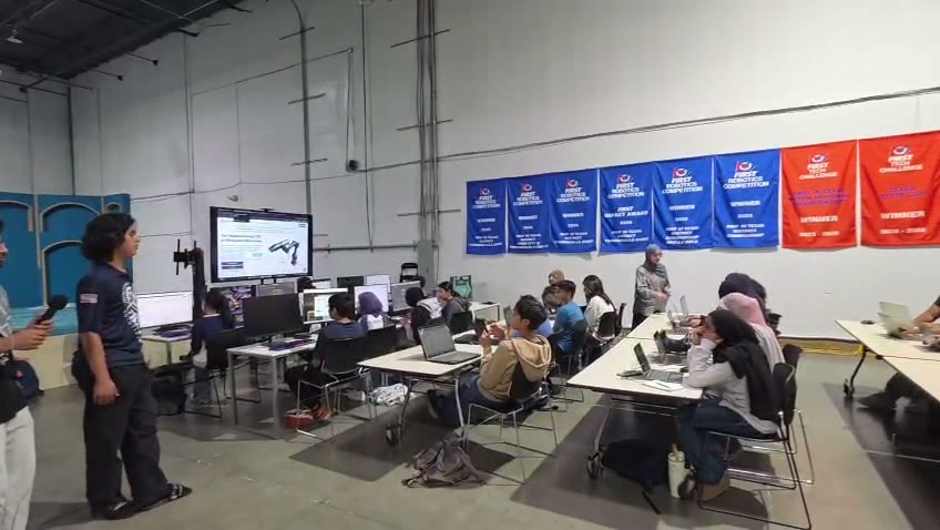 ITKAN students at laptops during a CAD training session, a CAD model on the big screen beneath the team's FIRST banner wall
